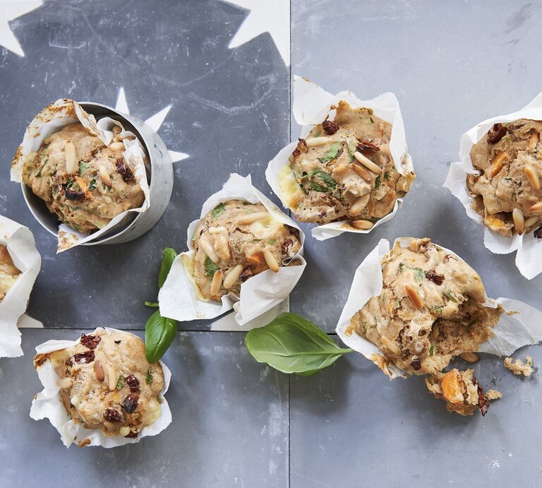 Vegetarische Gemüse-Pizza-Muffins mit Pinienkernen und frischem Basilikum, eingewickelt in Backpapier und drappiert auf einem hellen Metalltisch.  | © StockFood / Gräfe & Unzer Verlag / Brinkop, Maria