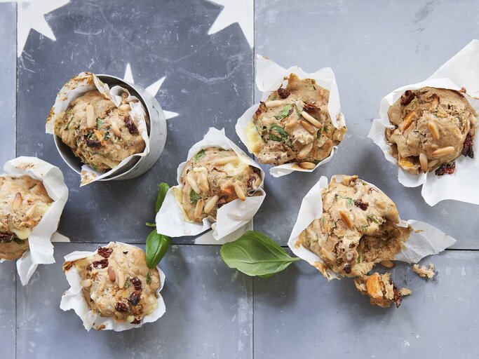 Vegetarische Gemüse-Pizza-Muffins mit Pinienkernen und frischem Basilikum, eingewickelt in Backpapier und drappiert auf einem hellen Metalltisch.  | © StockFood / Gräfe & Unzer Verlag / Brinkop, Maria