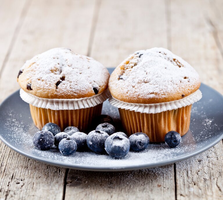 Frontalsicht: Auf einem Holzuntergrund steht ein grauer Teller mit zwei Joghurt-Muffins mit Beeren. Neben den Muffins liegen frische Blaubeeren. | © Shutterstock / Maria Kovalets