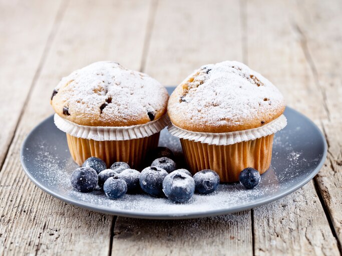 Frontalsicht: Auf einem Holzuntergrund steht ein grauer Teller mit zwei Joghurt-Muffins mit Beeren. Neben den Muffins liegen frische Blaubeeren. | © Shutterstock / Maria Kovalets
