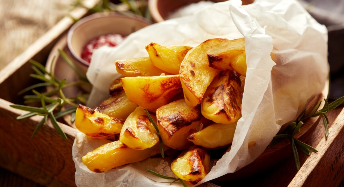 Eine Portion Kohlrabi-Pommes, eingewickelt in eine Papiertüte, auf einem Holztablett. Daneben Schälchen mit Dips.