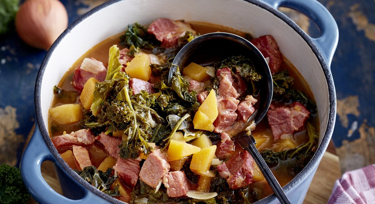 Kasseler-Gulasch in einem großen Topf mit Grünkohl, Kartoffeln und gewürfeltem Kasseler auf einer Tischplatte mit den einzelnen Zutaten drum herum. 