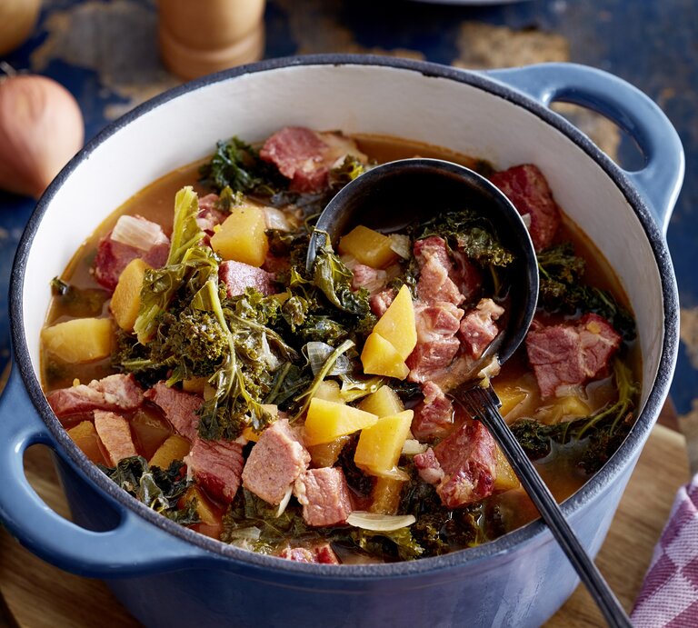 Kasseler-Gulasch in einem großen Topf mit Grünkohl, Kartoffeln und gewürfeltem Kasseler auf einer Tischplatte mit den einzelnen Zutaten drum herum.  | © EAT CLUB / Isabell Triemer