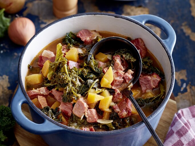 Kasseler-Gulasch in einem großen Topf mit Grünkohl, Kartoffeln und gewürfeltem Kasseler auf einer Tischplatte mit den einzelnen Zutaten drum herum.  | © EAT CLUB / Isabell Triemer
