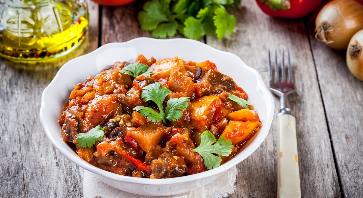 Eine Schale Kürbis-Kartoffelgulasch, drumherum liegen frischen Gemüse und Kräuter, außerdem eine Gabel.
