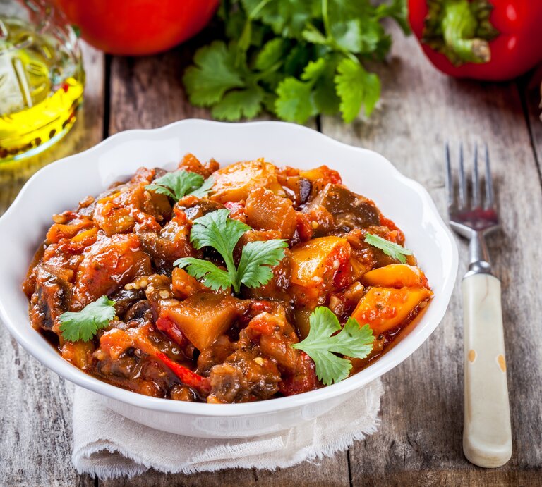 Eine Schale Kürbis-Kartoffelgulasch, drumherum liegen frischen Gemüse und Kräuter, außerdem eine Gabel. | © Adobe Stock/ nblxer