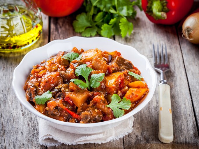 Eine Schale Kürbis-Kartoffelgulasch, drumherum liegen frischen Gemüse und Kräuter, außerdem eine Gabel. | © Adobe Stock/ nblxer
