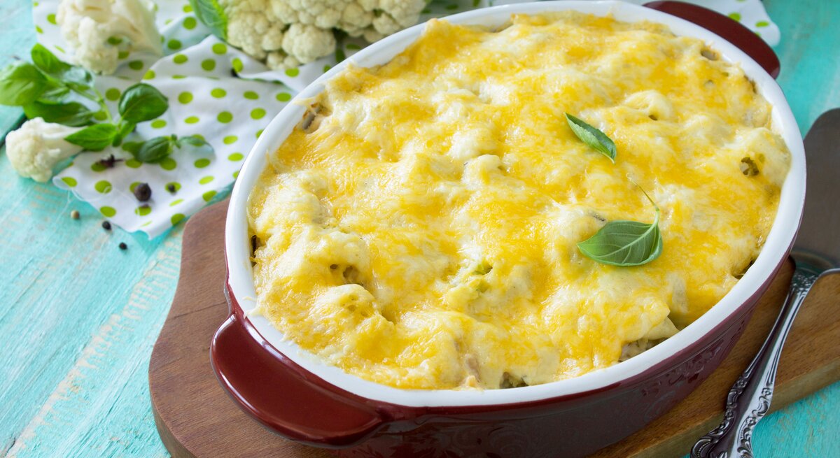 Draufsicht, schräg: Auf einem hellblauen Untergrund aus Holz steht eine ovale Ofenform mit einem Blumenkohl-Lachs-Auflauf mit Gouda auf einem Brettchen. Daneben liegt roher Blumenkohl.