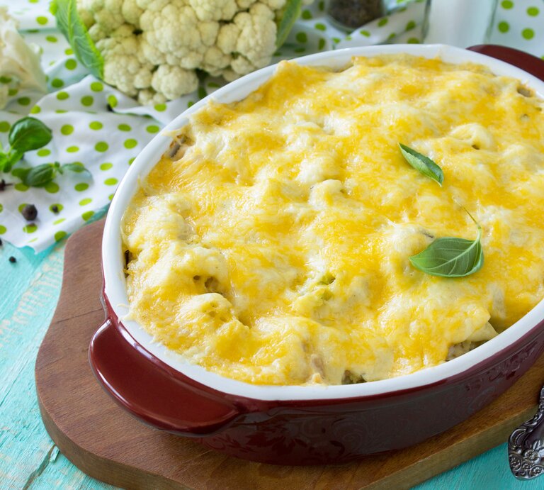 Draufsicht, schräg: Auf einem hellblauen Untergrund aus Holz steht eine ovale Ofenform mit einem Blumenkohl-Lachs-Auflauf mit Gouda auf einem Brettchen. Daneben liegt roher Blumenkohl. | © Shutterstock / Elena Hramova