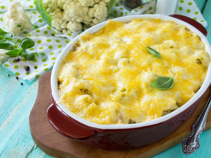 Draufsicht, schräg: Auf einem hellblauen Untergrund aus Holz steht eine ovale Ofenform mit einem Blumenkohl-Lachs-Auflauf mit Gouda auf einem Brettchen. Daneben liegt roher Blumenkohl. | © Shutterstock / Elena Hramova