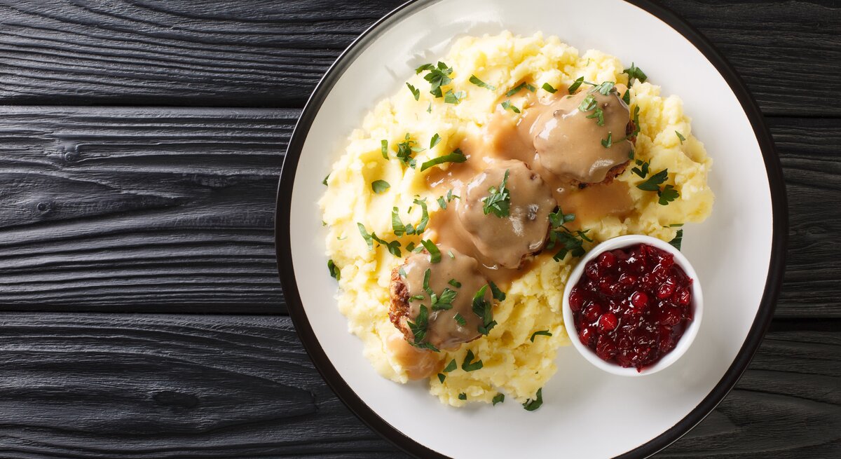 Von-Oben-Ansicht eines Tellers mit finnischen Hackbällchen, cremiger Soße und Kartoffelbrei. Daneben steht eine Schale Kompott. 