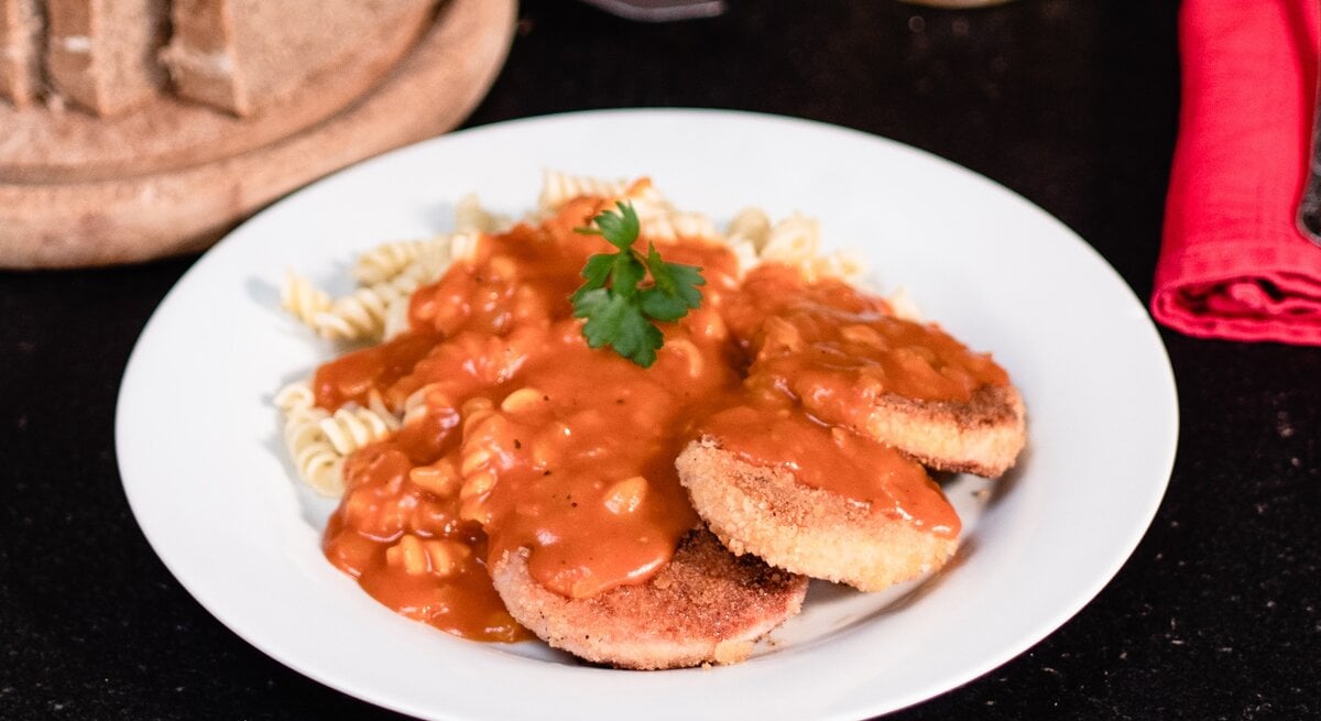 DDR Jägerschnitzel – panierte Jagdwurstscheiben mit Spirelli-Nudeln Tomatensoße. Ostdeutsches Gericht auf weißem Teller. Daneben angeschnittenes Brot und Besteck. 