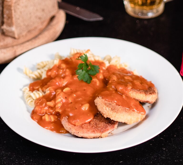 DDR Jägerschnitzel – panierte Jagdwurstscheiben mit Spirelli-Nudeln Tomatensoße. Ostdeutsches Gericht auf weißem Teller. Daneben angeschnittenes Brot und Besteck.  | © BILD der FRAU/Pako Cardenas Quijada