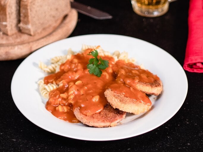 DDR Jägerschnitzel – panierte Jagdwurstscheiben mit Spirelli-Nudeln Tomatensoße. Ostdeutsches Gericht auf weißem Teller. Daneben angeschnittenes Brot und Besteck.  | © BILD der FRAU/Pako Cardenas Quijada