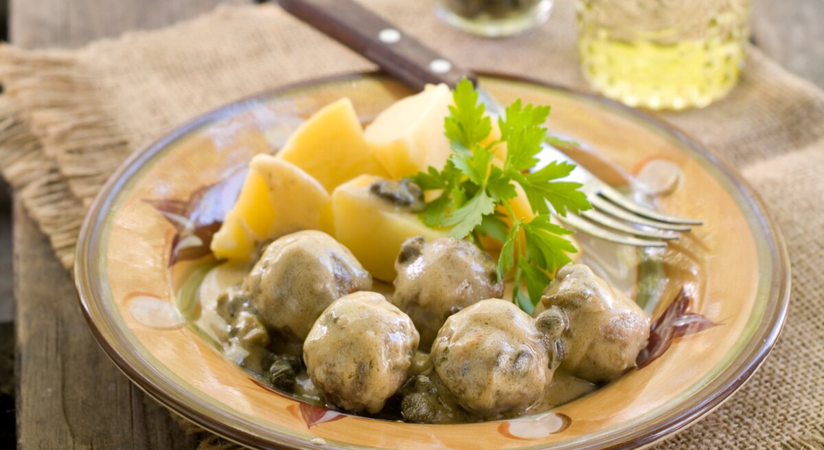 Königsberger Klopse mit Kapern und Soße zu Kartoffeln auf einem kitschigen Teller. 