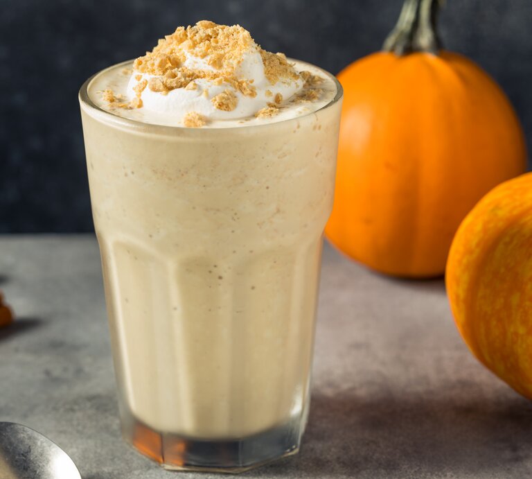Frontalsicht: Ein Glas mit einem Kürbis-Milkshake mit Vanilleeis steht auf einem dunklen Untergrund. Neben dem Glas liegen Zimtstangen, eine Löffel sowie zwei kleine Kürbisse. | © Shutterstock / bhofack2