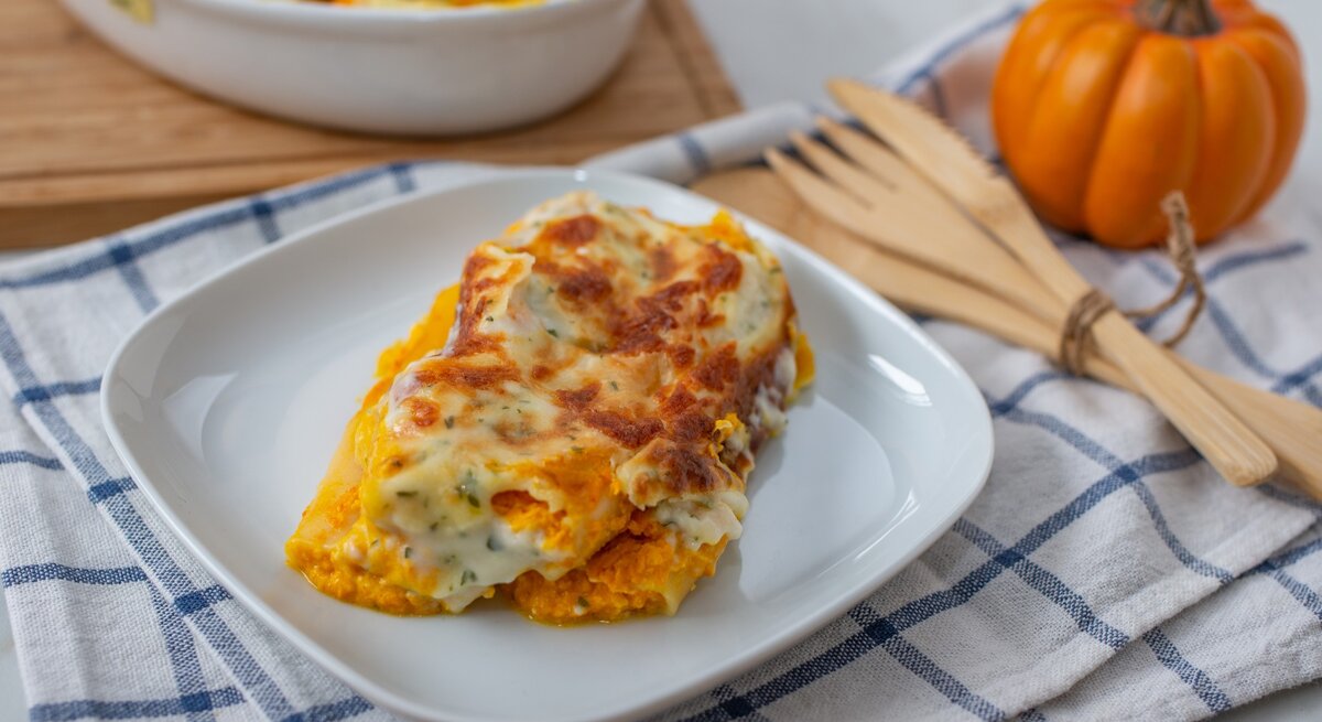 Kürbis-Cannelloni mit Kräuter-Bechamel und Mozzarella überbacken auf weißem Teller und blau-weißem Tuch aus der Draufsicht. Holzbesteck rechts mit Schnur zusammengebunden. Daneben kleiner Kürbis leicht verschwommen. Links oben angedeutete weiße Auflaufform auf Holzbrett.