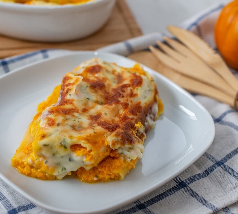 Kürbis-Cannelloni mit Kräuter-Bechamel und Mozzarella überbacken auf weißem Teller und blau-weißem Tuch aus der Draufsicht. Holzbesteck rechts mit Schnur zusammengebunden. Daneben kleiner Kürbis leicht verschwommen. Links oben angedeutete weiße Auflaufform auf Holzbrett. | © Getty Images/A_lein