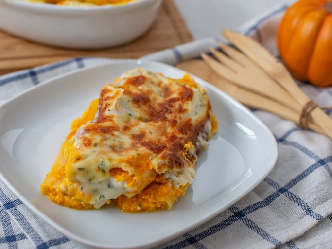 Kürbis-Cannelloni mit Kräuter-Bechamel und Mozzarella überbacken auf weißem Teller und blau-weißem Tuch aus der Draufsicht. Holzbesteck rechts mit Schnur zusammengebunden. Daneben kleiner Kürbis leicht verschwommen. Links oben angedeutete weiße Auflaufform auf Holzbrett. | © Getty Images/A_lein