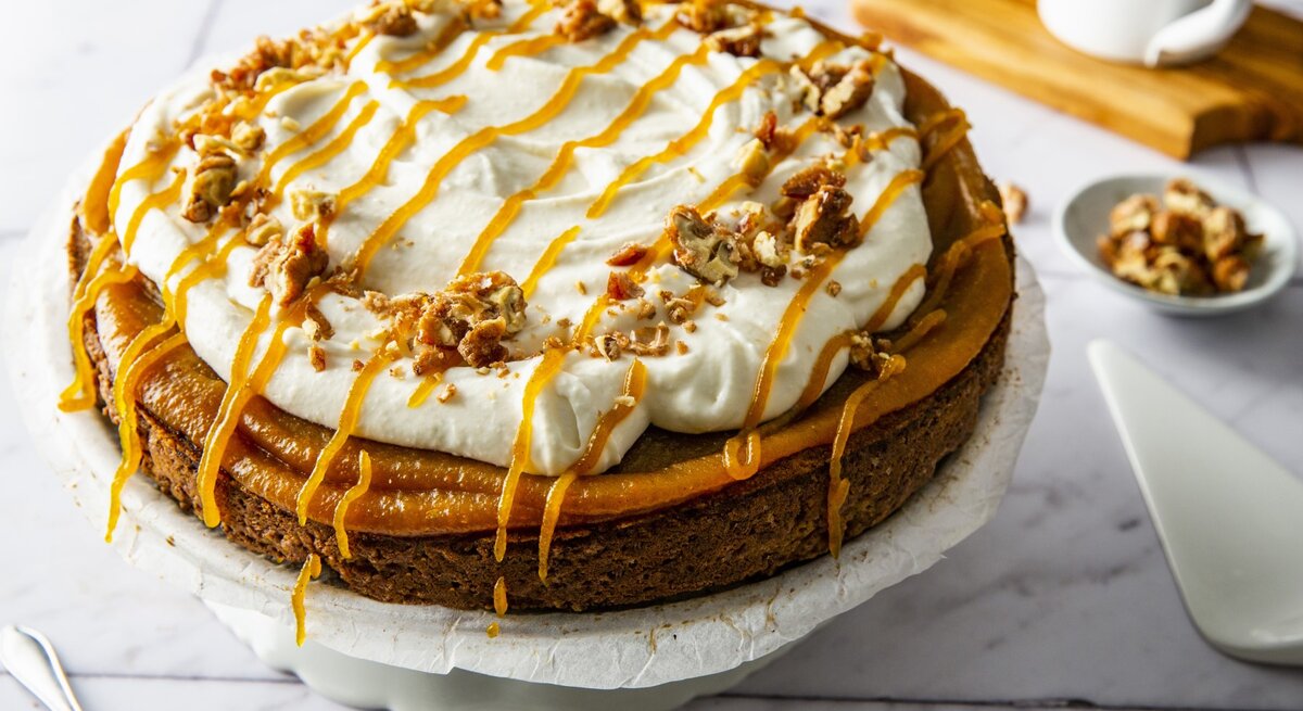 Kürbiskuchen mit Frischkäsecreme und gebrannten Walnüssen, garniert mit Karamell und serviert auf einer großen Kuchenplatte auf einem weißen Fliesentisch. 