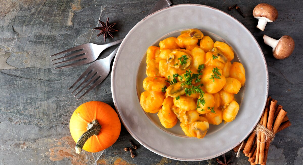 Eine Portion Kürbis-Gnocchi mit Kürbis-Pilz-Soße in einem weißen Teller steht auf einem Untergrund aus Stein. Um den Teller herum liegen Zimt, Sternanis, ein Kürbis, Pilze und zwei Gabeln.