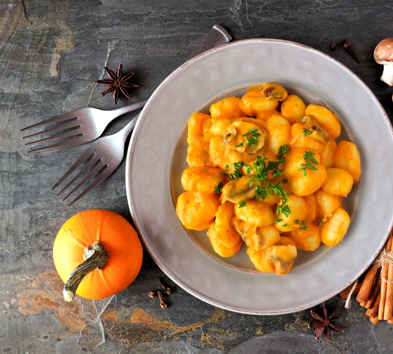 Eine Portion Kürbis-Gnocchi mit Kürbis-Pilz-Soße in einem weißen Teller steht auf einem Untergrund aus Stein. Um den Teller herum liegen Zimt, Sternanis, ein Kürbis, Pilze und zwei Gabeln. | © Getty Images / jenifoto