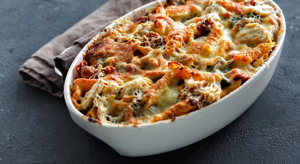 Kürbis-Nudelauflauf mit Spinat in einer ovalen Auflaufform, mit Käse überbacken, daneben ein graues Geschirrtuch.