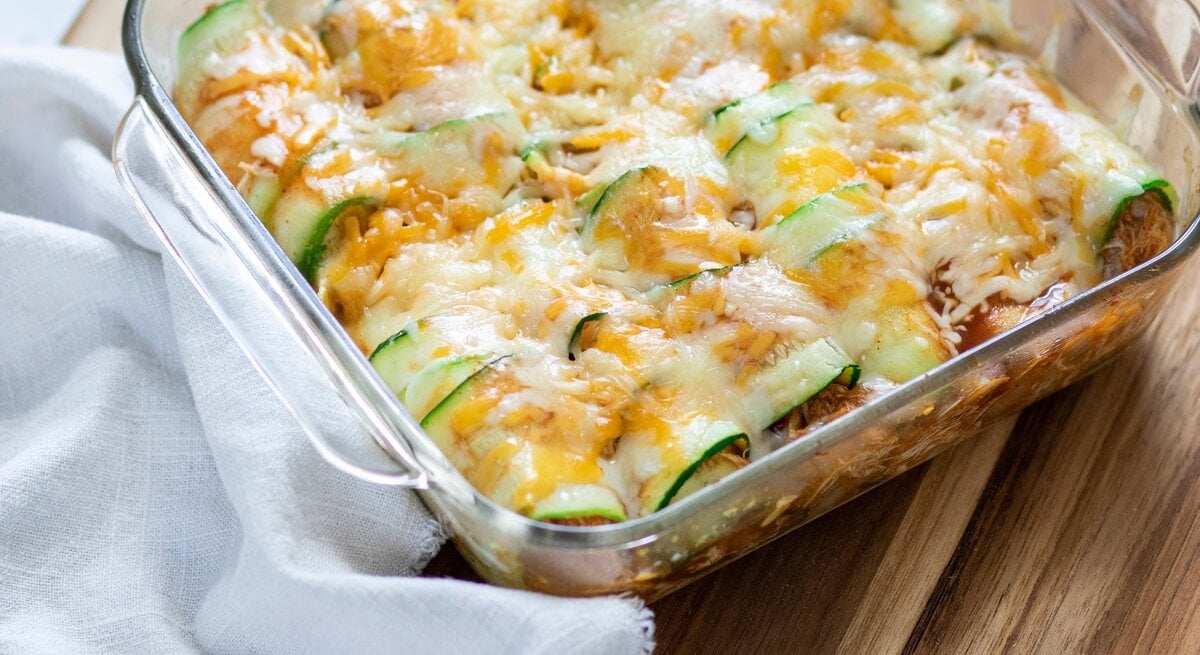 Überbackene Zucchini-Enchiladas mit Hack in einer Auflaufform. Daneben ein Tuch. Das Ganze auf einem hölzernen Untergrund. 