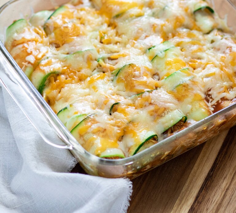 Überbackene Zucchini-Enchiladas mit Hack in einer Auflaufform. Daneben ein Tuch. Das Ganze auf einem hölzernen Untergrund.  | © Shutterstock/ Allison McAdams
