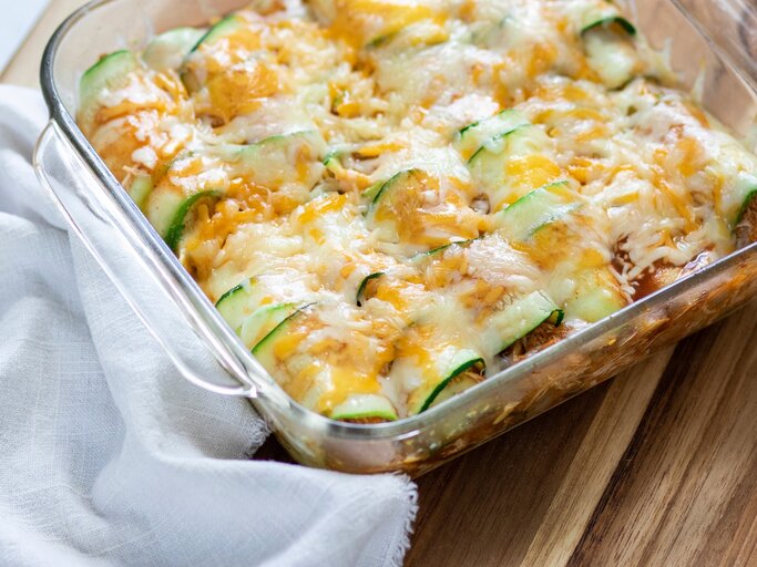 Überbackene Zucchini-Enchiladas mit Hack in einer Auflaufform. Daneben ein Tuch. Das Ganze auf einem hölzernen Untergrund.  | © Shutterstock/ Allison McAdams