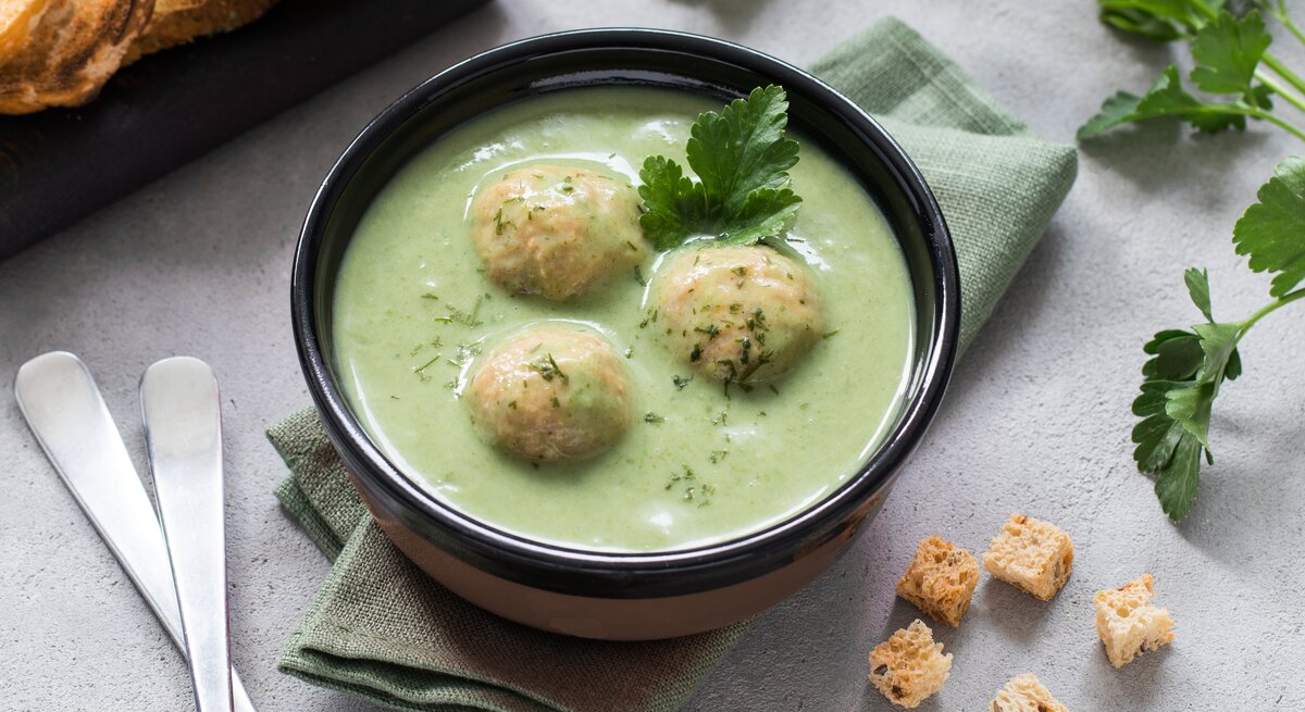 Zucchinisuppe mit Hackfleisch-Bällchen in schwarzer Schüssel auf hellem Untergrund. Daneben Kräuter, Löffel und geröstetes Brot. 
