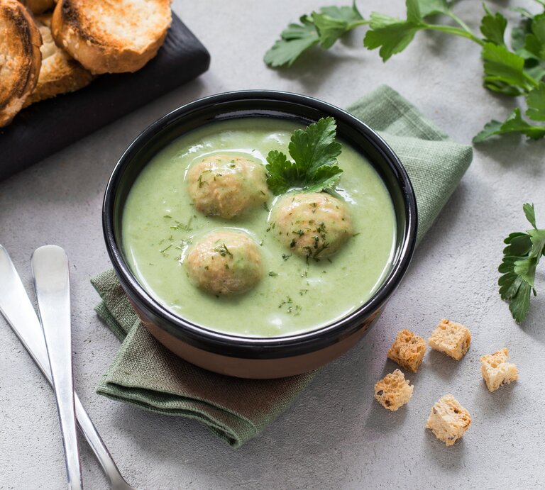 Zucchinisuppe mit Hackfleisch-Bällchen in schwarzer Schüssel auf hellem Untergrund. Daneben Kräuter, Löffel und geröstetes Brot.  | © Shutterstock/Aksana Ban