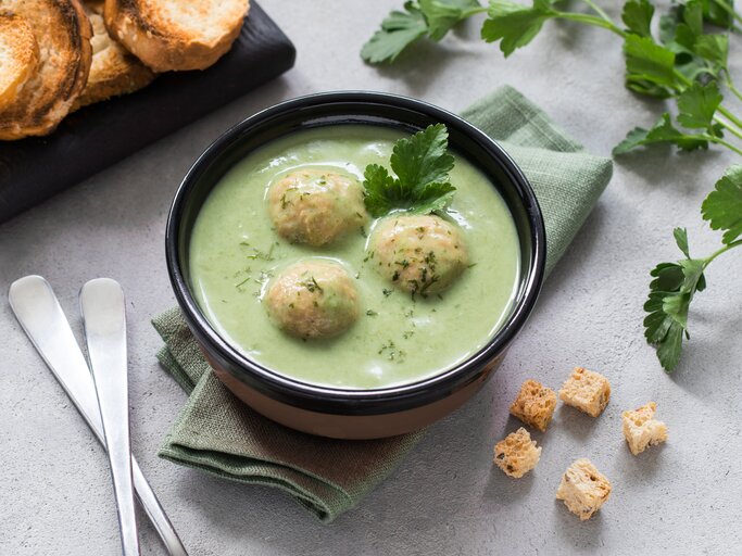Zucchinisuppe mit Hackfleisch-Bällchen in schwarzer Schüssel auf hellem Untergrund. Daneben Kräuter, Löffel und geröstetes Brot.  | © Shutterstock/Aksana Ban