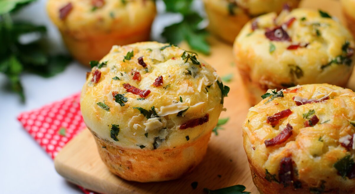 Einige Pizza-Muffins auf einem Holzschneidebrett und darunter eine rot-weiße Decke. Heller Hintergrund und einige Kräuter sind auch noch zu sehen.