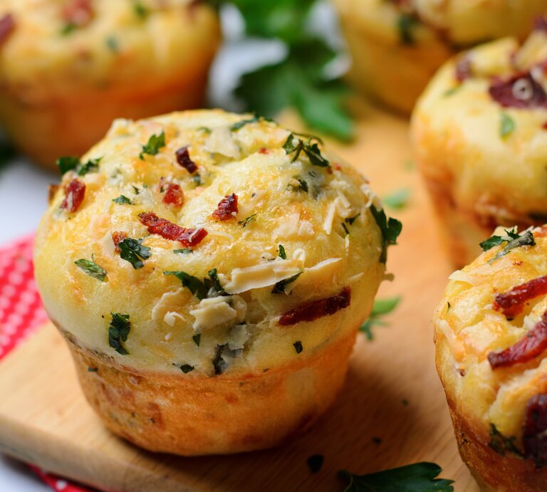 Einige Pizza-Muffins auf einem Holzschneidebrett und darunter eine rot-weiße Decke. Heller Hintergrund und einige Kräuter sind auch noch zu sehen. | © Shutterstock/ Julie208