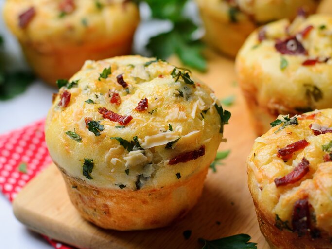 Einige Pizza-Muffins auf einem Holzschneidebrett und darunter eine rot-weiße Decke. Heller Hintergrund und einige Kräuter sind auch noch zu sehen. | © Shutterstock/ Julie208
