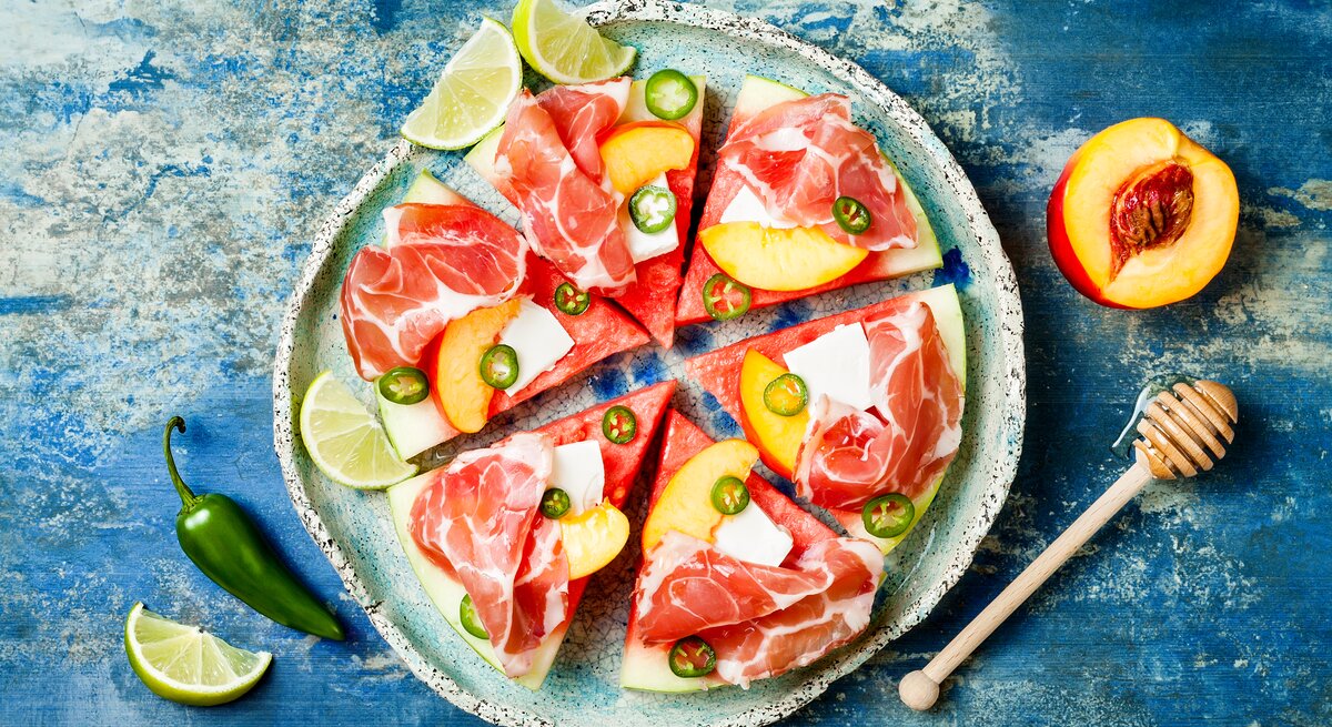 Eine Scheibe Wassermelone unterteilt in sechs gleichgroße Pizzastücke belegt mit Feta, Prosciutto, Jalapeños und Aprikosen, neben Limetten Stücken, von oben, vor einem blauen Hintergrund. 