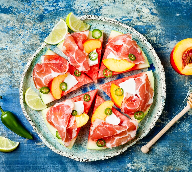 Eine Scheibe Wassermelone unterteilt in sechs gleichgroße Pizzastücke belegt mit Feta, Prosciutto, Jalapeños und Aprikosen, neben Limetten Stücken, von oben, vor einem blauen Hintergrund.  | © GettyImages/sveta_zarzamora