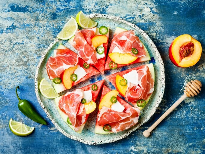 Eine Scheibe Wassermelone unterteilt in sechs gleichgroße Pizzastücke belegt mit Feta, Prosciutto, Jalapeños und Aprikosen, neben Limetten Stücken, von oben, vor einem blauen Hintergrund.  | © GettyImages/sveta_zarzamora