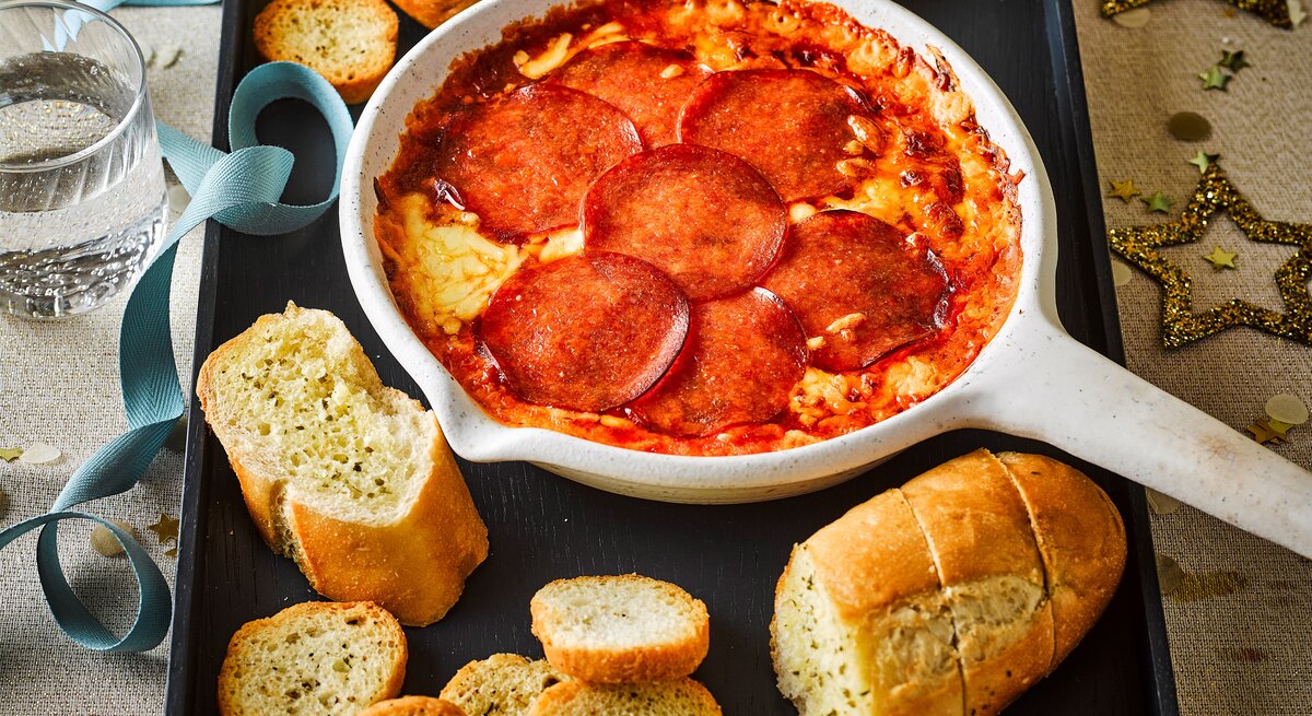Eine weiße, ofenfeste Pfanne mit geschmolzenem Käse und einer Schicht aus runden Salami-Scheiben gefüllt. Angerichtet als Pizza-Fondue-Dip. Draufsicht