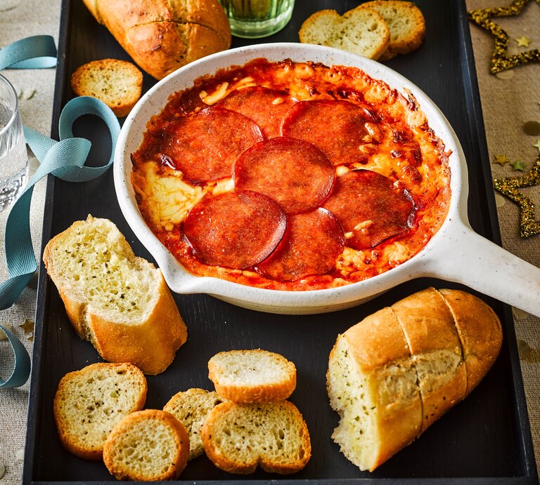 Eine weiße, ofenfeste Pfanne mit geschmolzenem Käse und einer Schicht aus runden Salami-Scheiben gefüllt. Angerichtet als Pizza-Fondue-Dip. Draufsicht | © StockFood / Immediate Media / Good Food