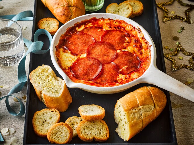 Eine weiße, ofenfeste Pfanne mit geschmolzenem Käse und einer Schicht aus runden Salami-Scheiben gefüllt. Angerichtet als Pizza-Fondue-Dip. Draufsicht | © StockFood / Immediate Media / Good Food