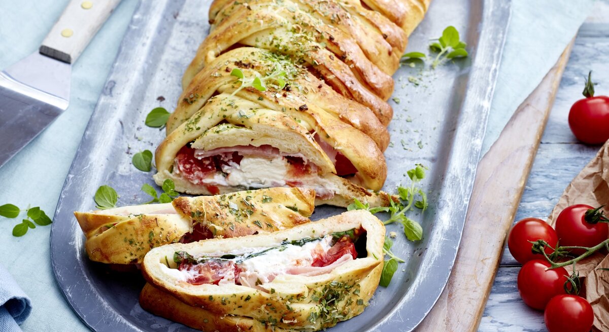 Auf einem Teller ist eine Pizzarolle „Stromboli“ angerichtet. Sie ist mit Aufschnitt gefüllt und mit Kräutern verfeinert.