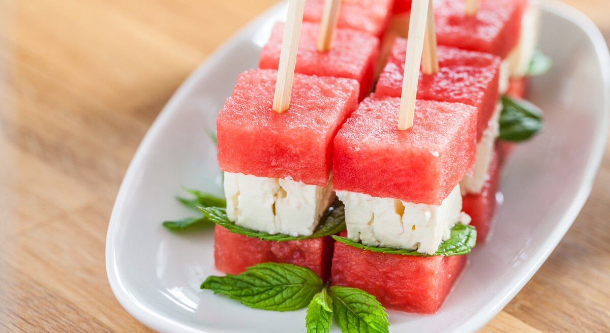 Wassermelonen-Feta-Spieße auf weißem Teller, angerichtet auf hölzernem Untergrund. 
