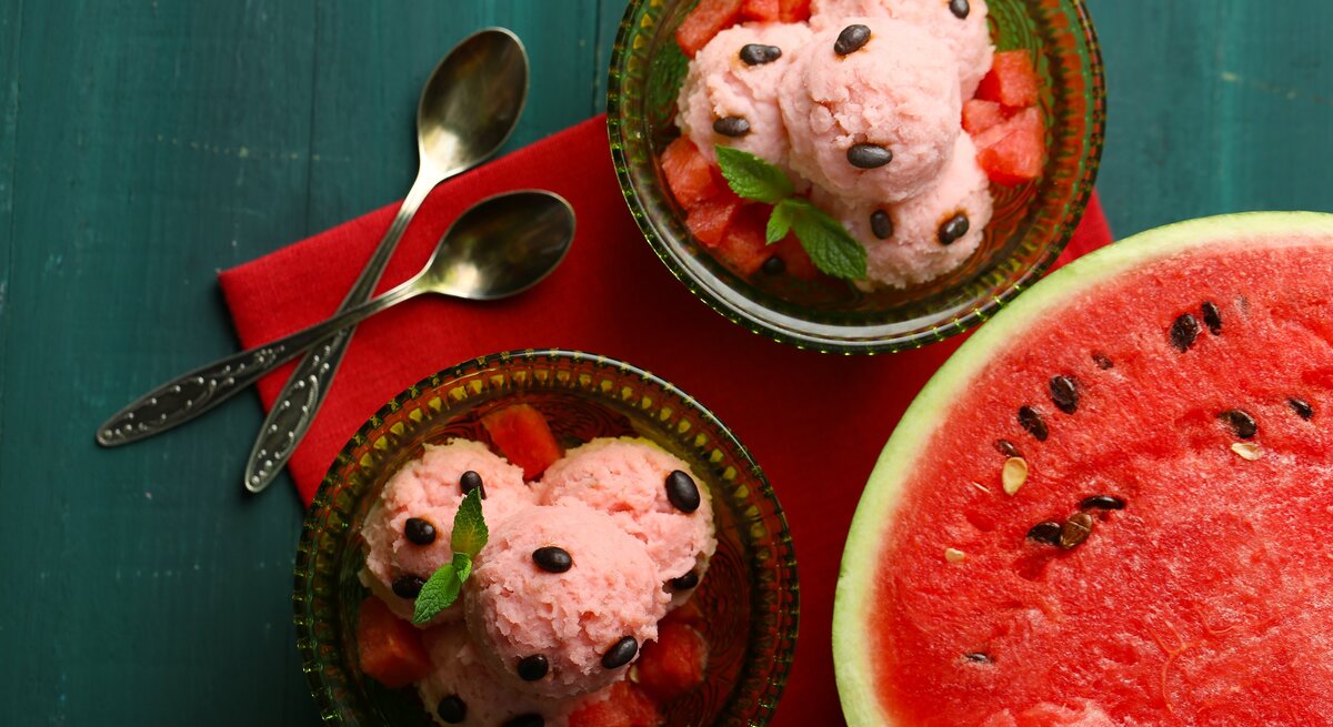 Draufsicht: Dunkelblauer Untergrund, zwei Schälchen aus Glas mit selbst gemachten Wassermelonen-Eis, dass auf einem bett von wassermelonen-Würfeln liegt und mit Wassermelonen-Kernen und einem Minzblatt dekoriert ist. Daneben liegt eine aufgeschnittene Wassermelonen-Hälfte.
