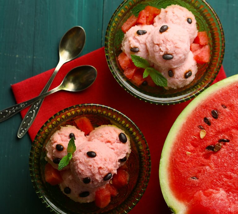 Draufsicht: Dunkelblauer Untergrund, zwei Schälchen aus Glas mit selbst gemachten Wassermelonen-Eis, dass auf einem bett von wassermelonen-Würfeln liegt und mit Wassermelonen-Kernen und einem Minzblatt dekoriert ist. Daneben liegt eine aufgeschnittene Wassermelonen-Hälfte. | © Schutterstock / Africa Studio