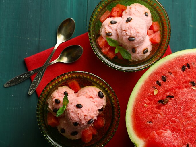 Draufsicht: Dunkelblauer Untergrund, zwei Schälchen aus Glas mit selbst gemachten Wassermelonen-Eis, dass auf einem bett von wassermelonen-Würfeln liegt und mit Wassermelonen-Kernen und einem Minzblatt dekoriert ist. Daneben liegt eine aufgeschnittene Wassermelonen-Hälfte. | © Schutterstock / Africa Studio