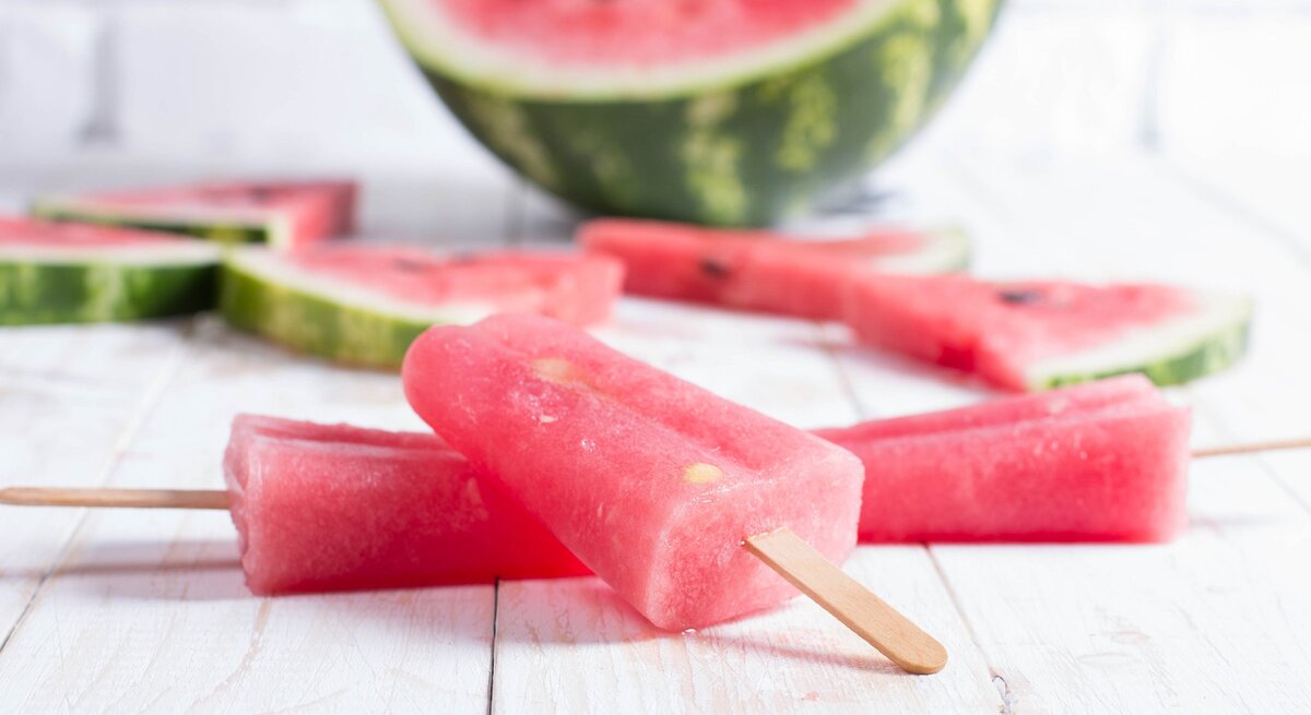 Ein Aperol Eis am Stiel, Stücke einer Wassermelone und eine halbierte Wasserlemone auf einem Untergrund aus Holz.