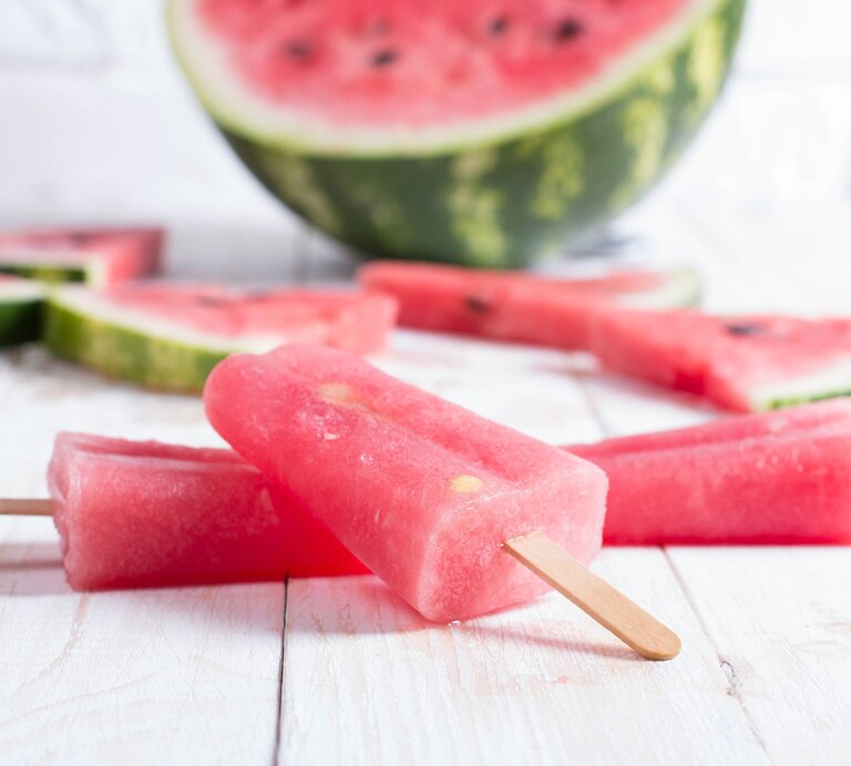 Ein Aperol Eis am Stiel, Stücke einer Wassermelone und eine halbierte Wasserlemone auf einem Untergrund aus Holz. | © GettyImages/ADragan
