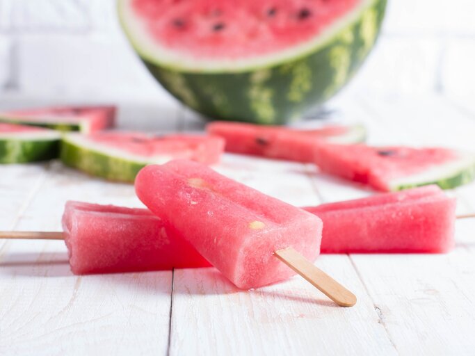 Ein Aperol Eis am Stiel, Stücke einer Wassermelone und eine halbierte Wasserlemone auf einem Untergrund aus Holz. | © GettyImages/ADragan